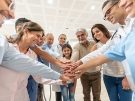 Multigenerational group of adults and a child standing in a circle indoors, smiling and stacking their hands together in a gesture of teamwork and support.